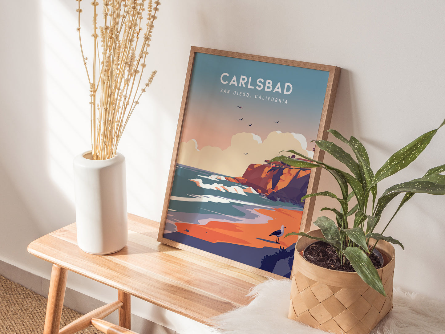 A framed Carlsbad, California poster rests on a wooden bench beside a white vase with dried plants and a potted green plant, creating a cozy.