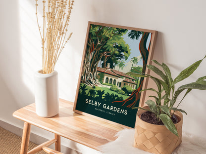 A framed Selby Gardens poster leans against a wall on a wooden bench beside a white vase with dried flowers and a potted plant.