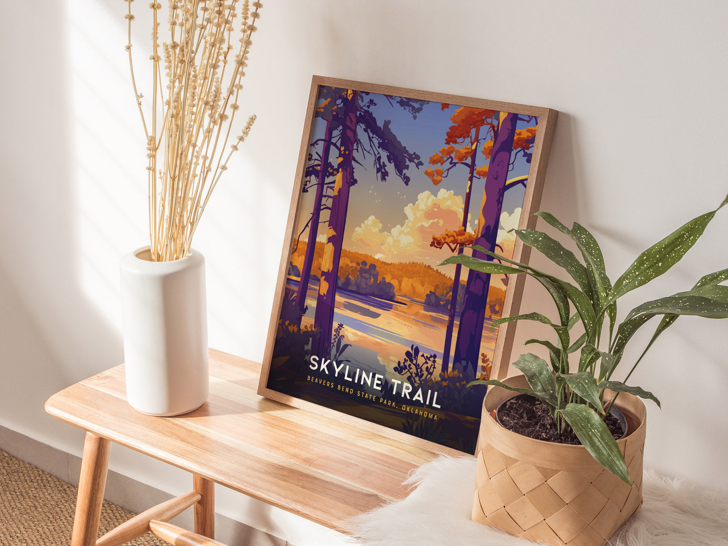 A framed Skyline Trail poster with vibrant colors leans against a wall on a wooden bench, accompanied by a white vase with dried branches and a potted plant.