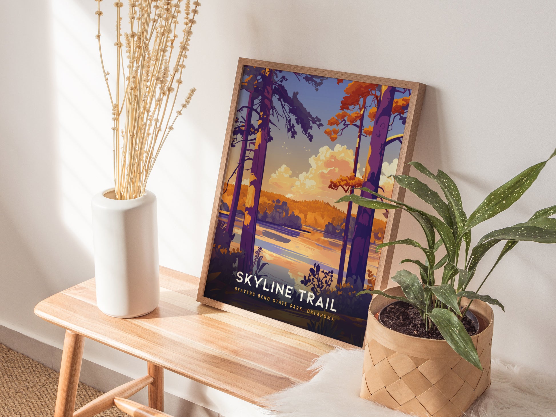 A framed Skyline Trail poster with vibrant colors leans against a wall on a wooden bench, accompanied by a white vase with dried branches and a potted plant.