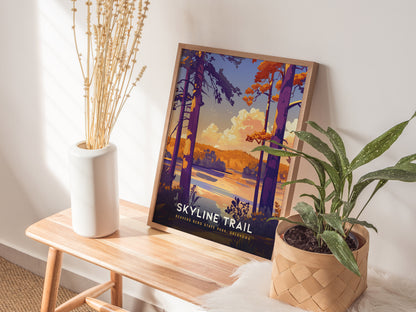 A framed Skyline Trail poster with vibrant colors leans against a wall on a wooden bench, accompanied by a white vase with dried branches and a potted plant.