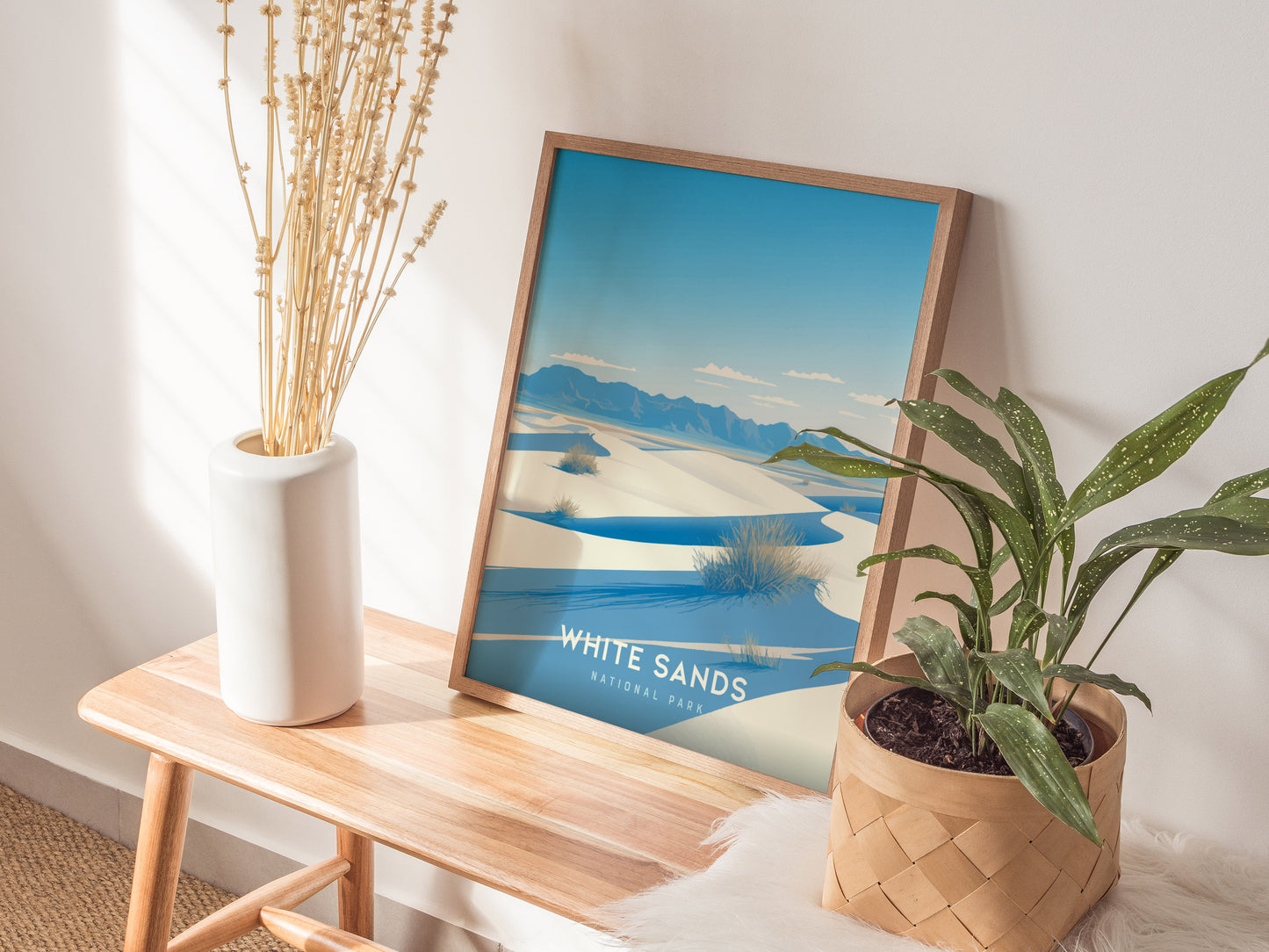A framed poster of White Sands National Park featuring desert dunes is displayed on a wooden bench, accompanied by a white vase with dried stems and a potted green plant.