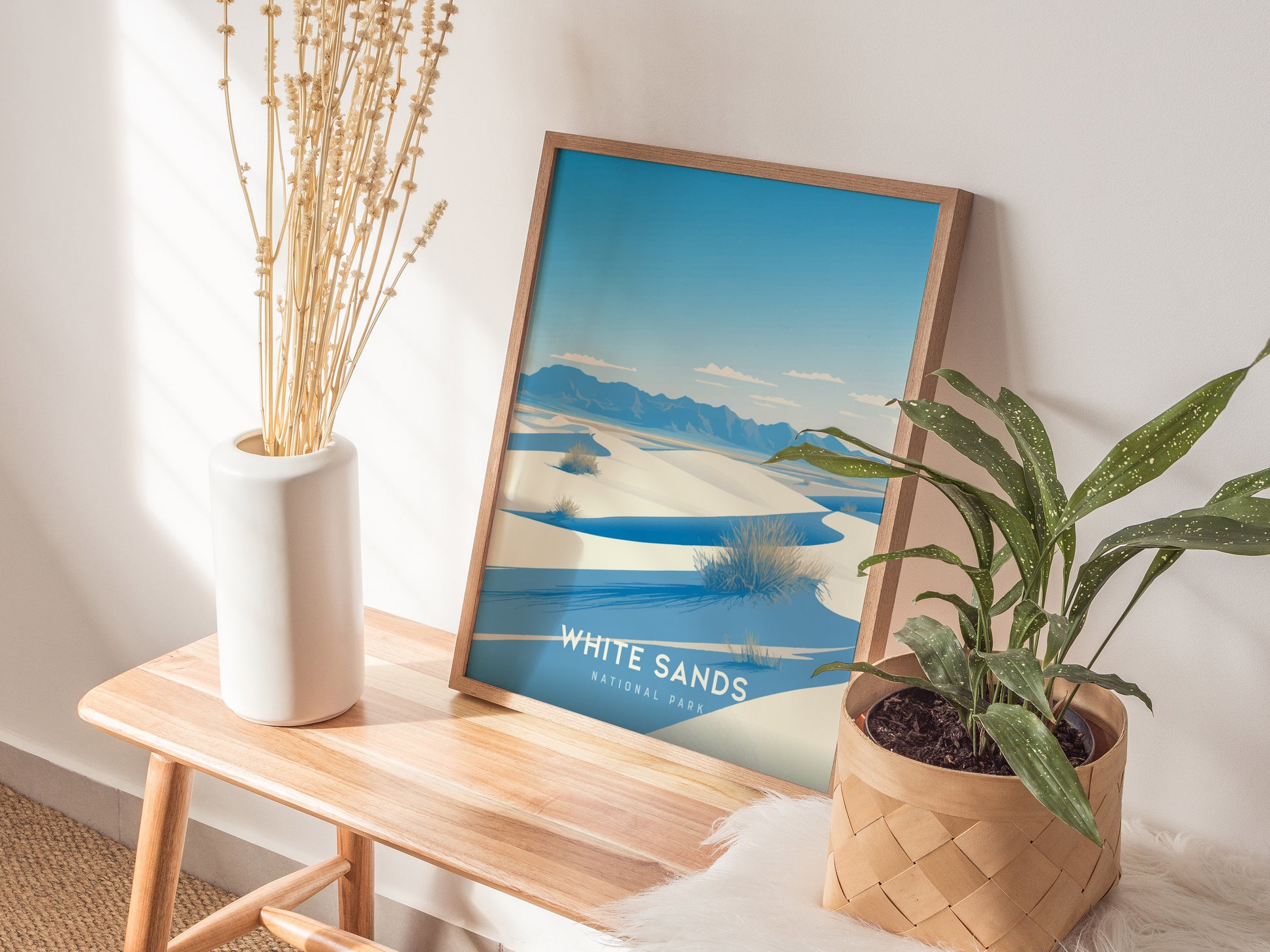 A framed poster of White Sands National Park featuring desert dunes is displayed on a wooden bench, accompanied by a white vase with dried stems and a potted green plant.