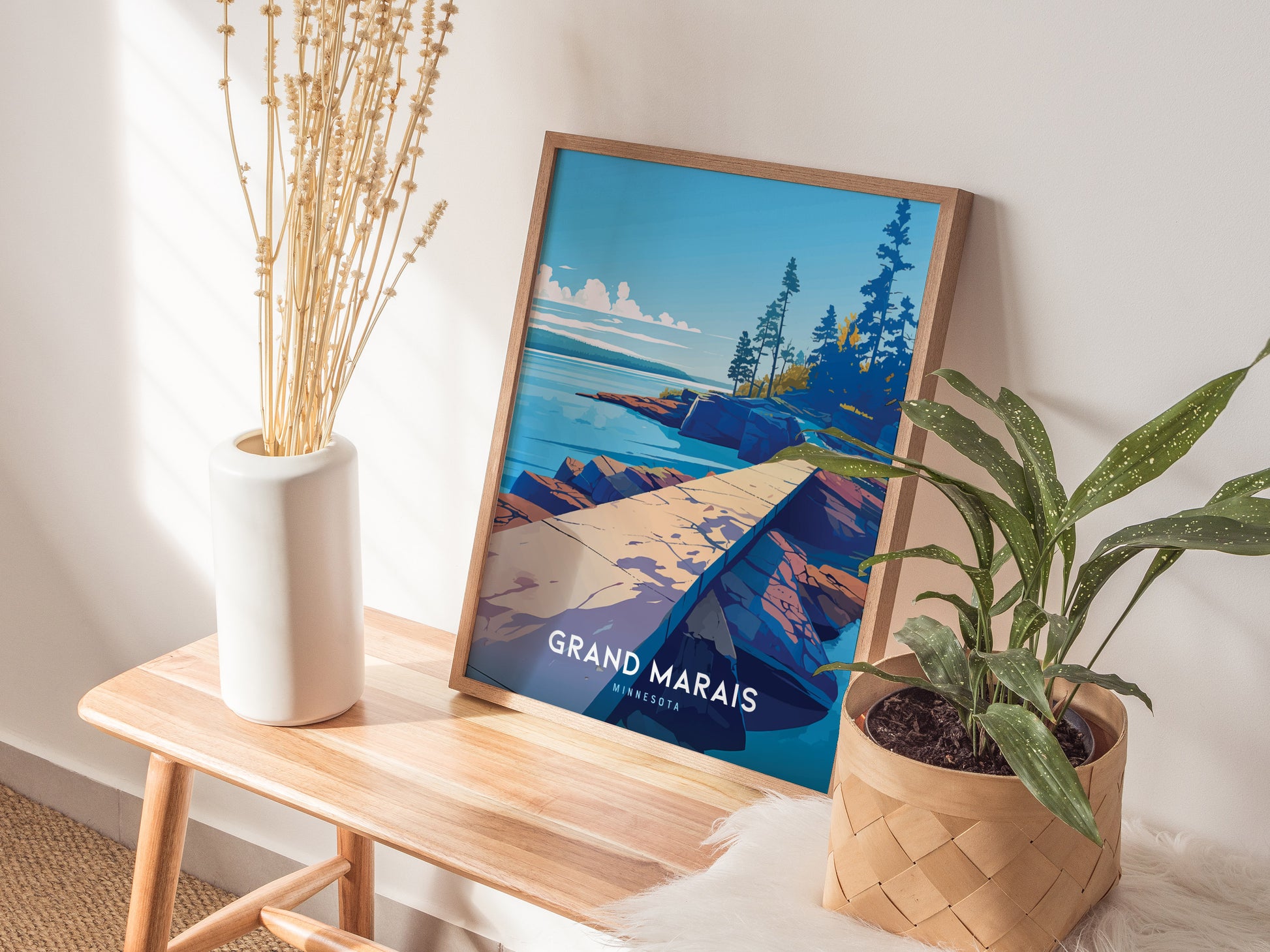 A framed Grand Marais poster featuring a scenic landscape rests on a wooden bench next to a white vase with dried flowers and a potted plant.