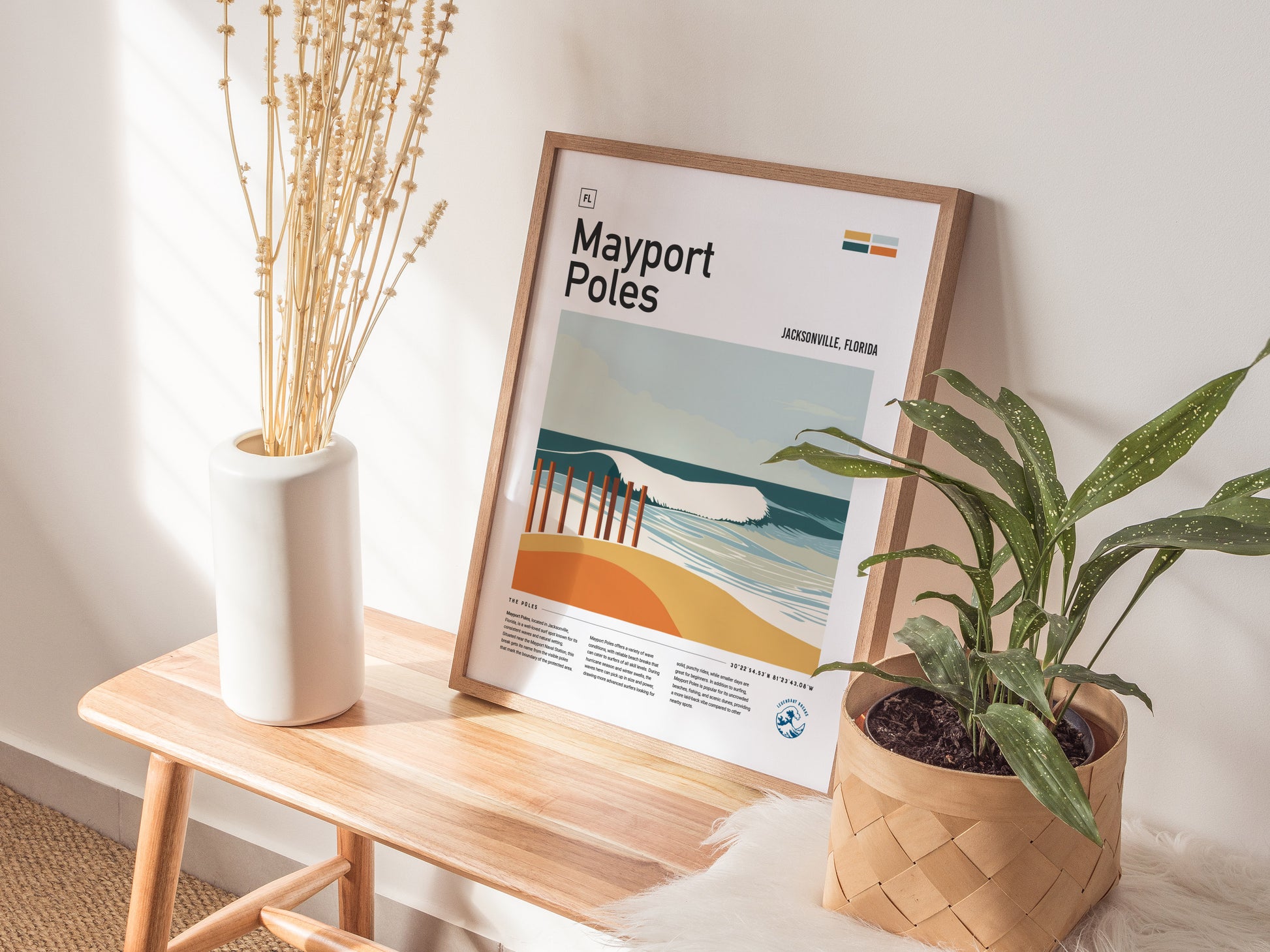 A framed Mayport Poles poster rests on a wooden bench, flanked by a white vase with dried flowers and a potted plant. Sunlight casts soft shadows on the wall.
