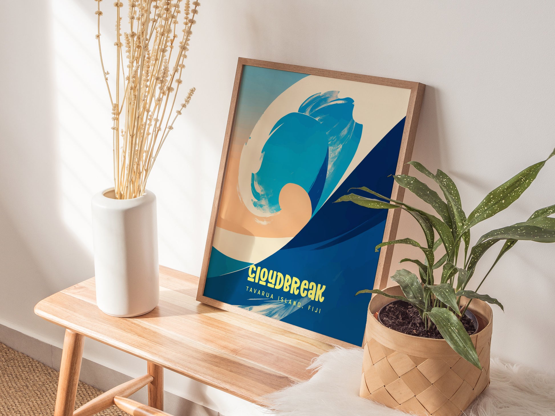 A surf-themed poster with blue and beige abstract wave art leans on a wooden bench, accompanied by a white vase with dried flowers and a green potted plant.