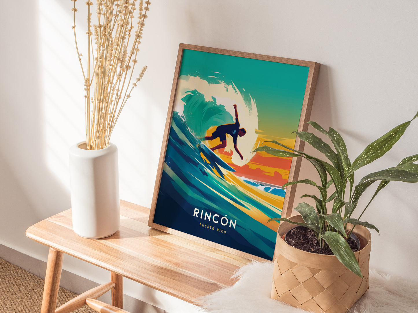 A framed surf poster titled Rincón Puerto Rico rests on a wooden table with a vase of dried flowers and a potted plant nearby, in a sunlit room.