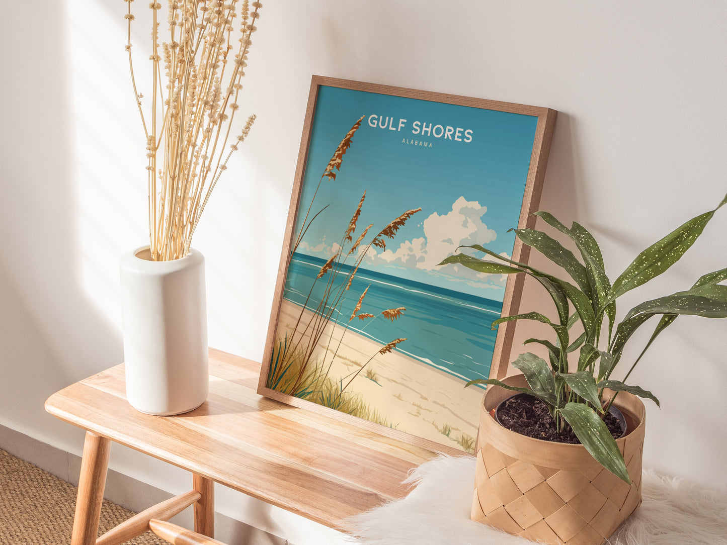 A framed Gulf Shores beach poster sits on a wooden bench beside a white vase with dried flowers and a potted green plant, creating a serene coastal vibe.