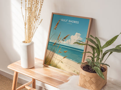 A framed Gulf Shores beach poster sits on a wooden bench beside a white vase with dried flowers and a potted green plant, creating a serene coastal vibe.