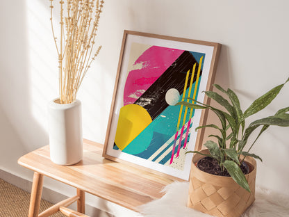 A framed abstract poster with vibrant 1980s-style geometric shapes and bold colors rests on a wooden bench, flanked by a white vase with dried stems and a potted plant.