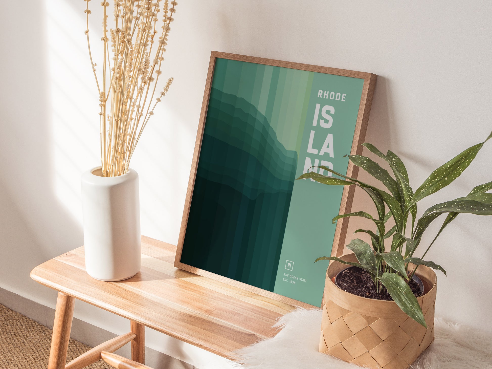 A framed abstract poster titled Rhode Island rests on a wooden bench, accompanied by a white vase with dried flowers and a potted plant, creating a modern decor setup.