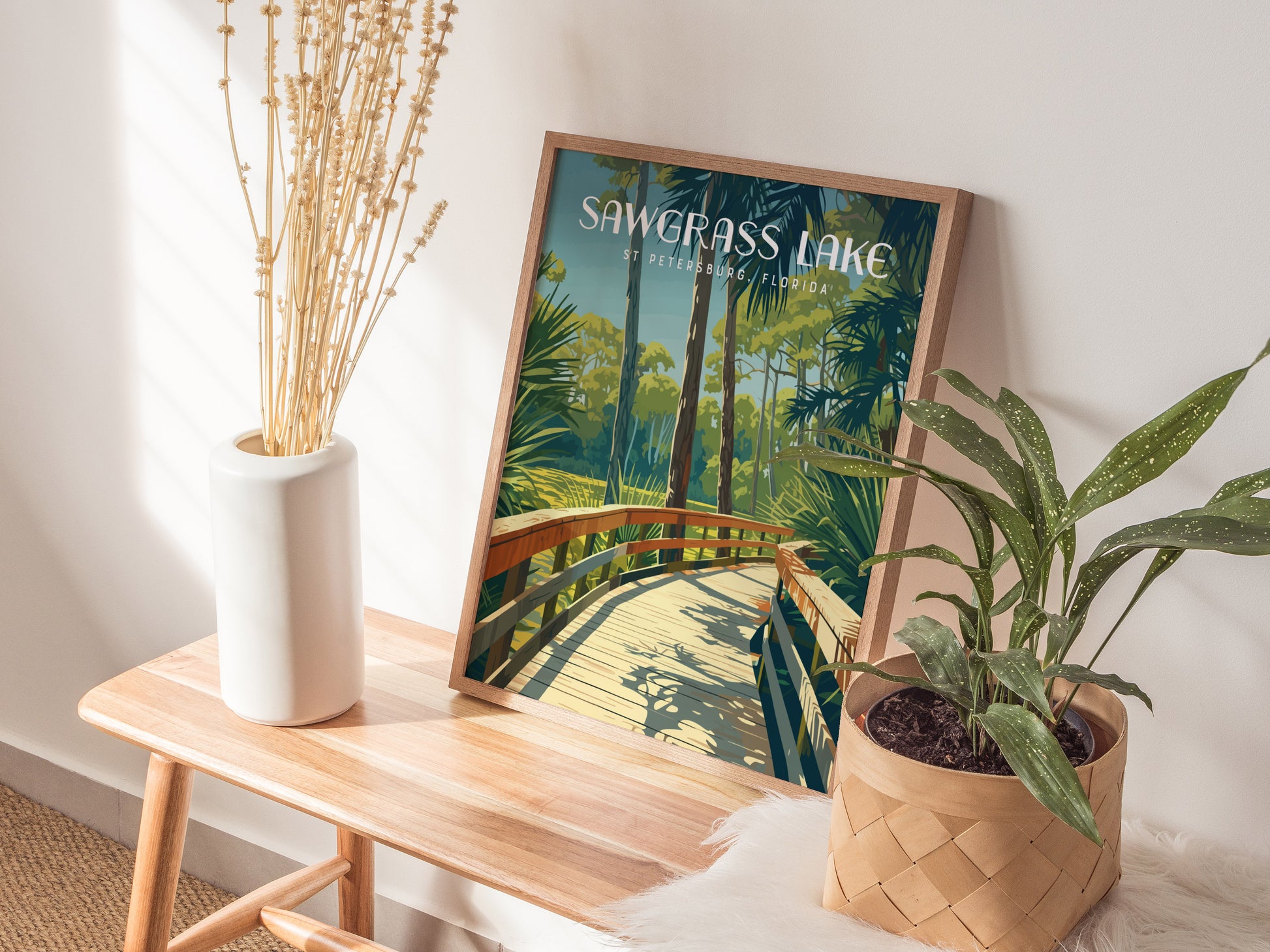 A framed Sawgrass Lake Park poster featuring a wooden boardwalk and lush greenery is displayed on a wooden bench with a white vase and a potted plant.