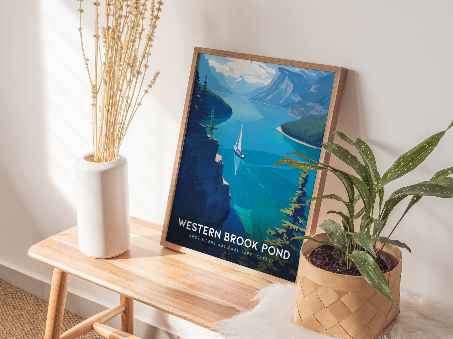 A framed poster of Western Brook Pond showing a fjord with a sailboat is placed on a wooden bench next to a white vase and a potted plant.