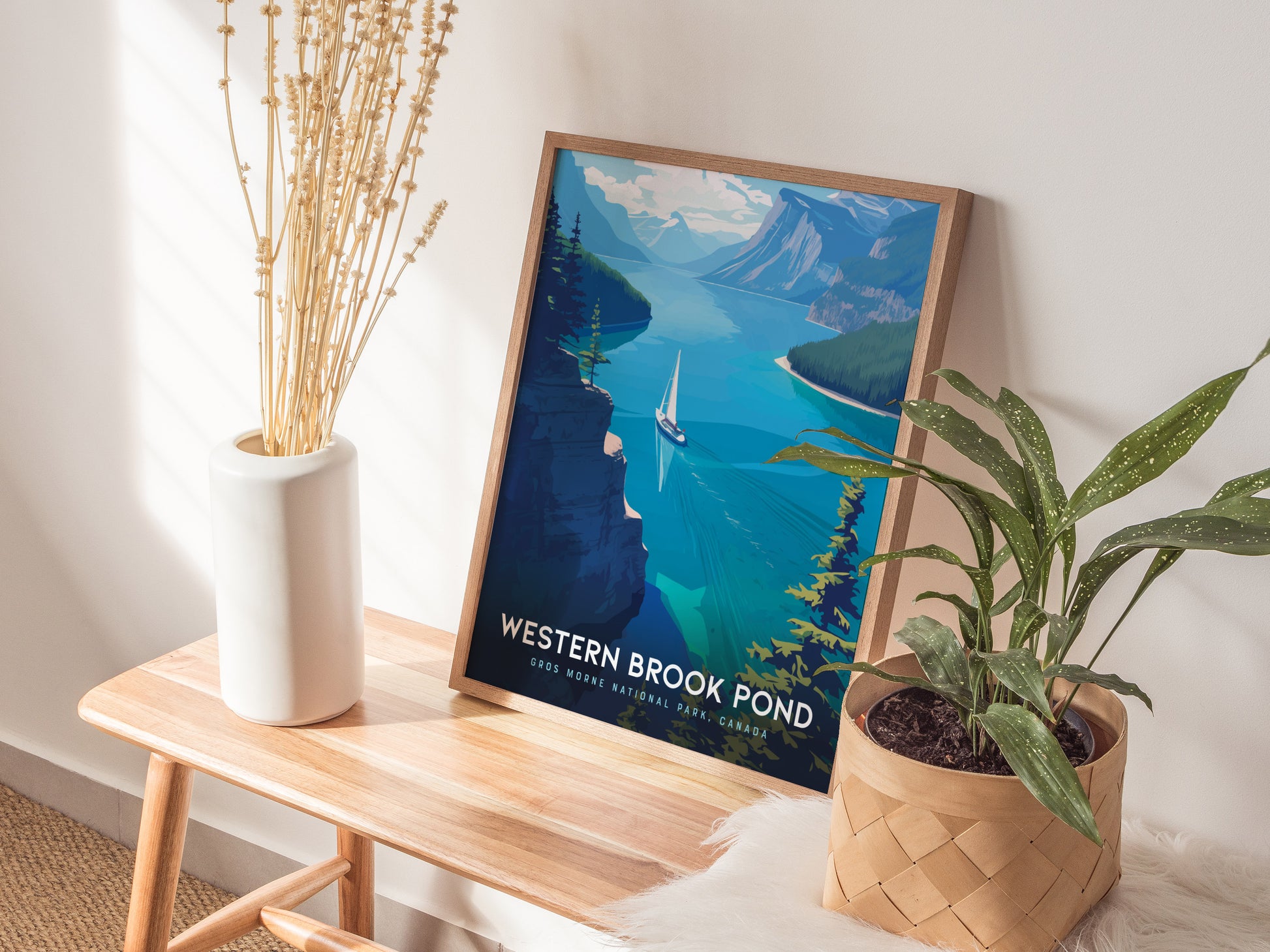 A framed poster of Western Brook Pond showing a fjord with a sailboat is placed on a wooden bench next to a white vase and a potted plant.
