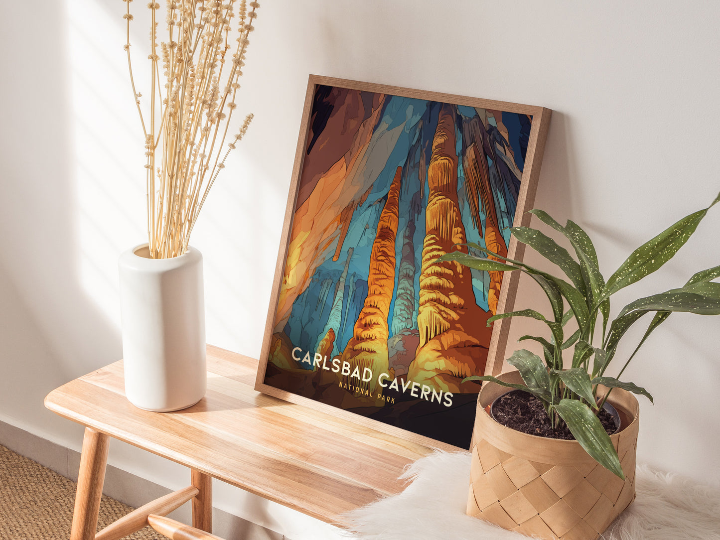 A Carlsbad Caverns National Park poster leans against a wall on a wooden bench, accompanied by a white vase with dried flowers and a potted green plant.