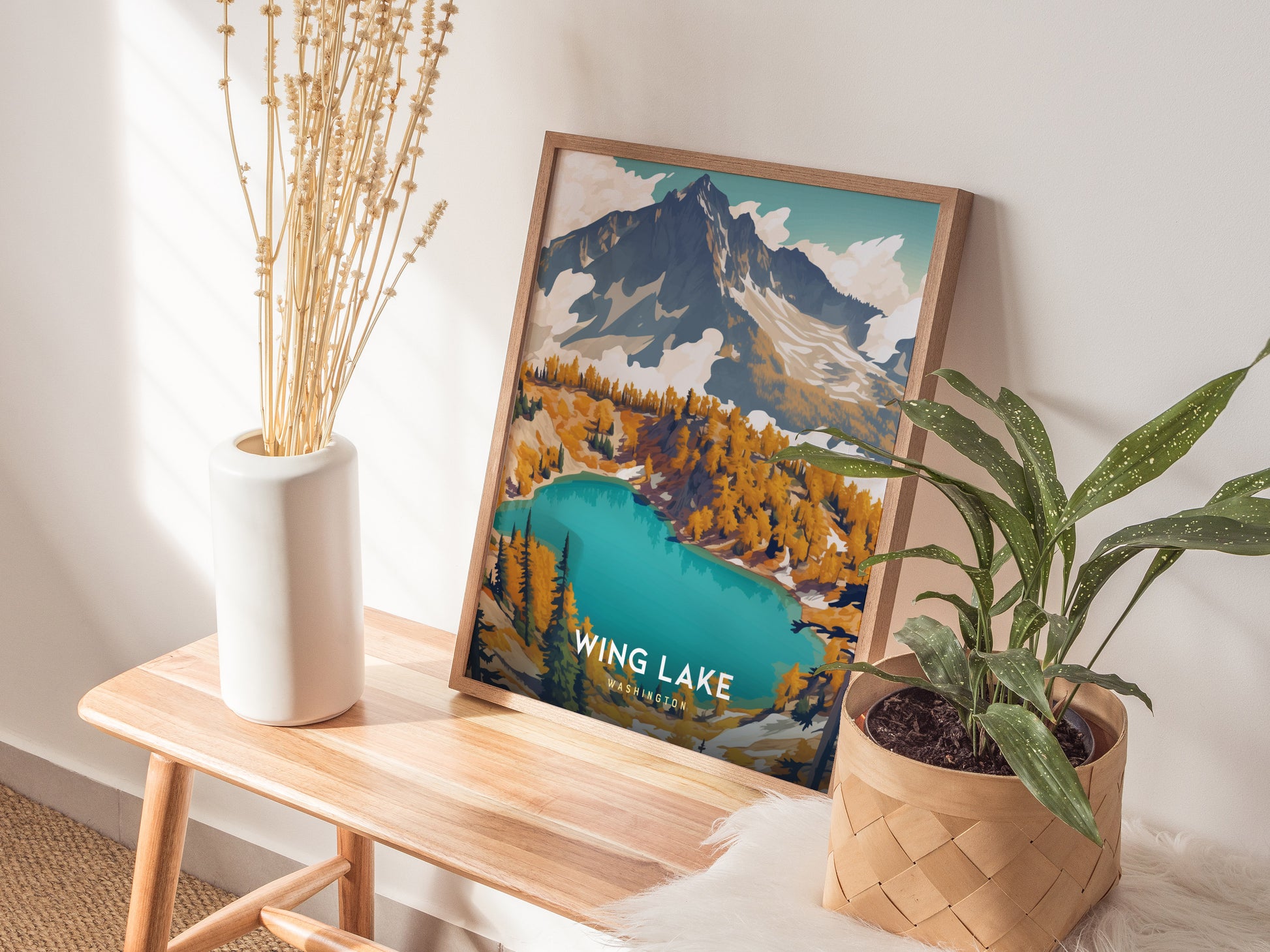 A framed Wing Lake Washington poster rests on a wooden bench, flanked by a white vase with dried flowers and a potted plant, in a sunlit room.