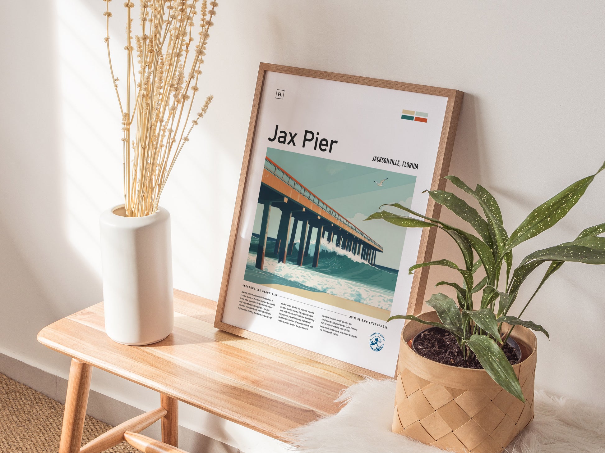 A framed Jax Pier surf poster rests on a wooden bench, accompanied by a white vase with dried stems and a potted plant, creating a cozy, natural decor setting.