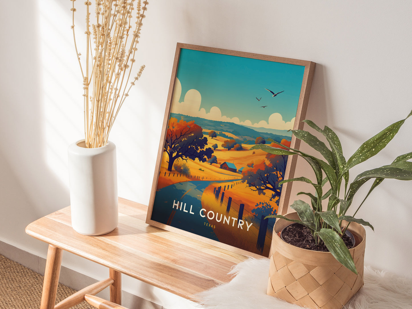 A framed Hill Country poster depicting vibrant Texas landscapes is placed on a wooden bench, accompanied by a white vase with dried stems and a potted plant.