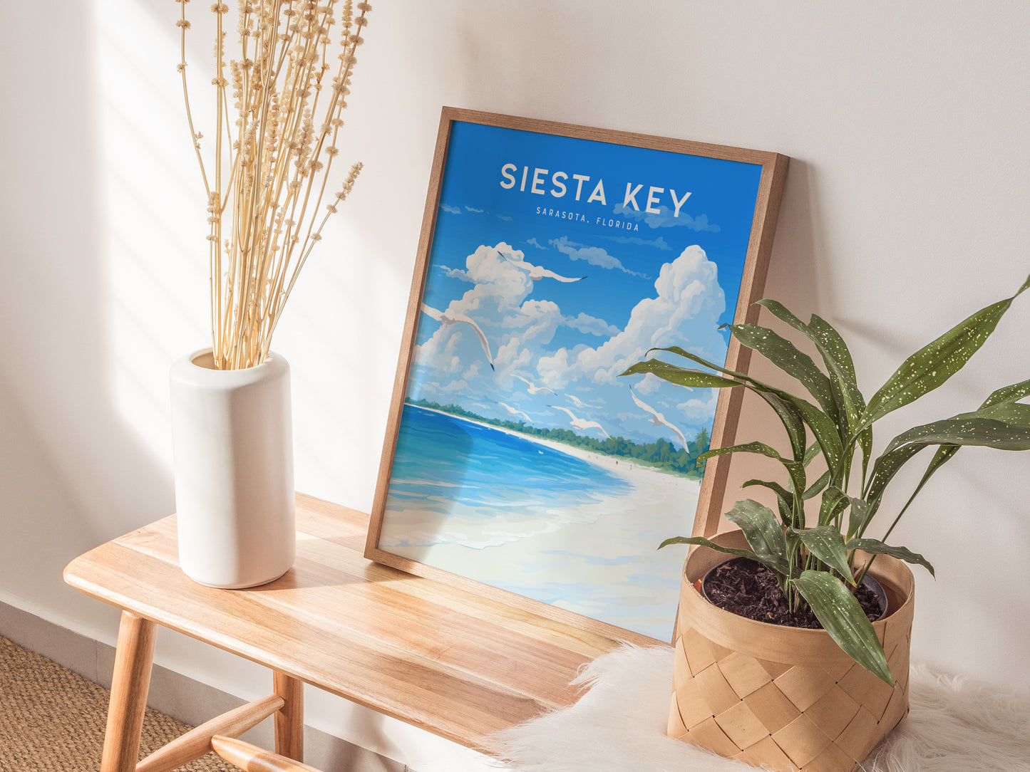 A framed Siesta Key poster depicting a beach scene with blue skies and white sand leans against a wall. Its placed on a wooden bench with a vase and a.