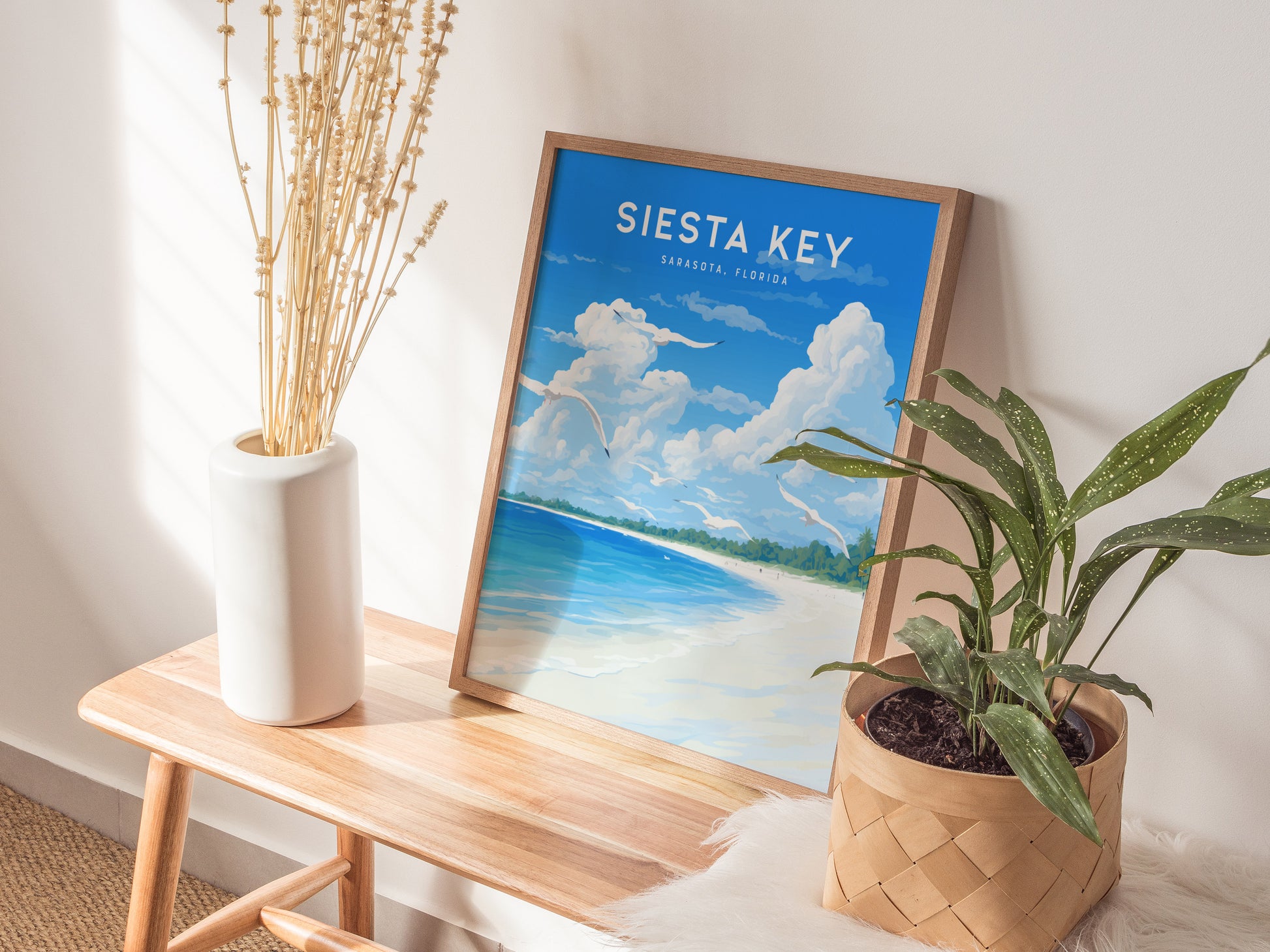 A framed Siesta Key poster depicting a beach scene with blue skies and white sand leans against a wall. Its placed on a wooden bench with a vase and a.