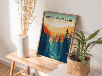 A framed Pacific Crest Trail poster with colorful forest imagery is placed on a wooden bench beside a white vase with dried flowers and a potted green plant.