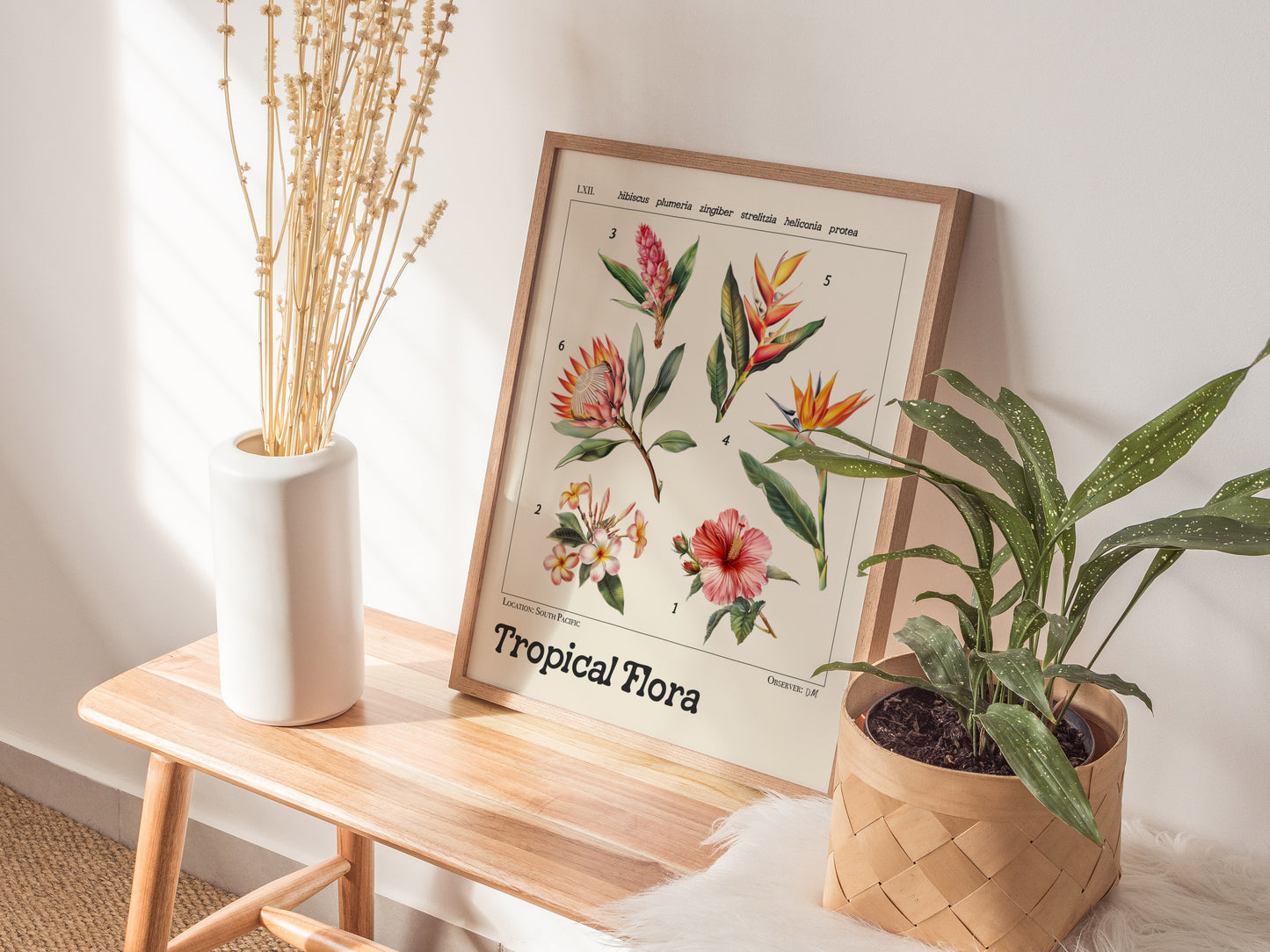 A framed botanical illustration poster featuring tropical flowers like hibiscus and bird of paradise is displayed on a wooden bench, alongside a white vase and a potted plant.