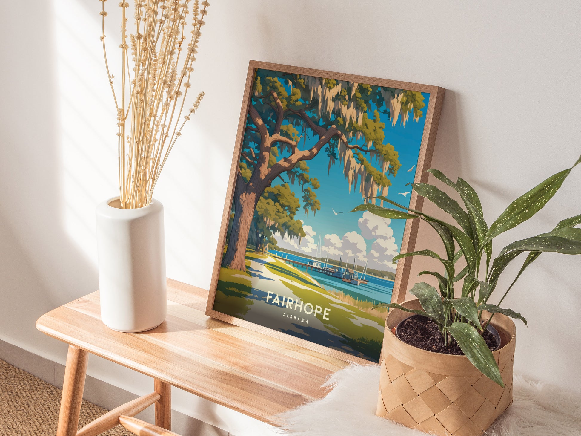 A framed poster of Fairhope, Alabama, depicting a coastal scene with a live oak tree, is displayed on a wooden bench next to a white vase and a potted plant.