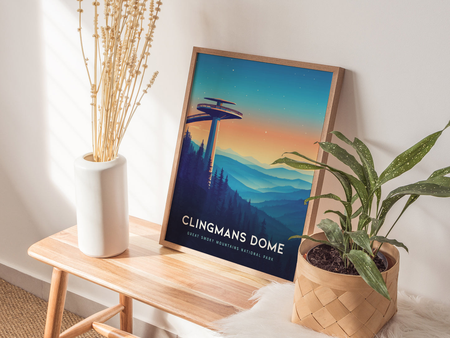 A framed Clingmans Dome poster rests on a wooden bench, depicting the observation tower against a colorful mountain landscape. The scene is complemented by a white vase with dry plants and a green potted plant.