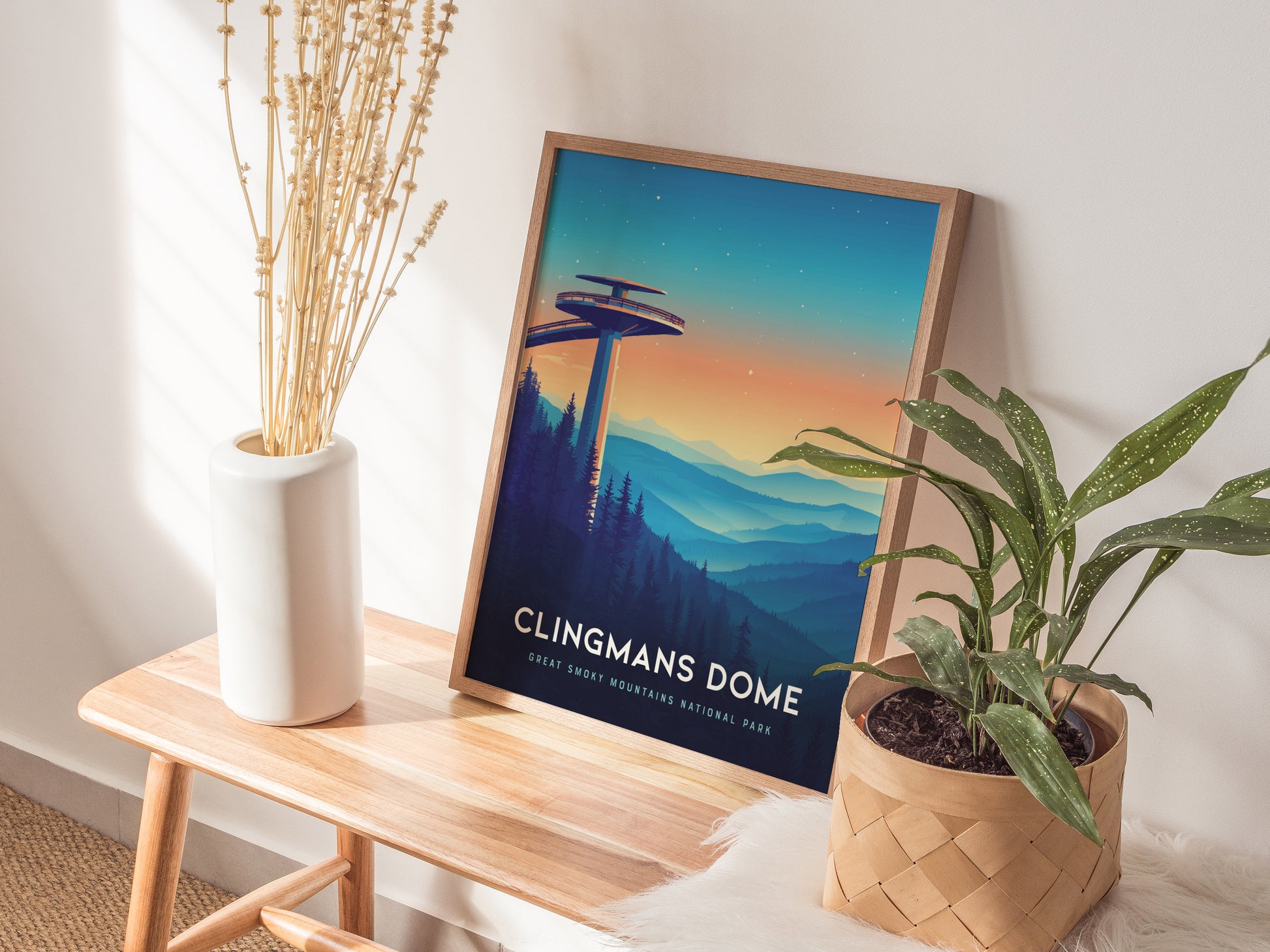 A framed Clingmans Dome poster rests on a wooden bench, depicting the observation tower against a colorful mountain landscape. The scene is complemented by a white vase with dry plants and a green potted plant.