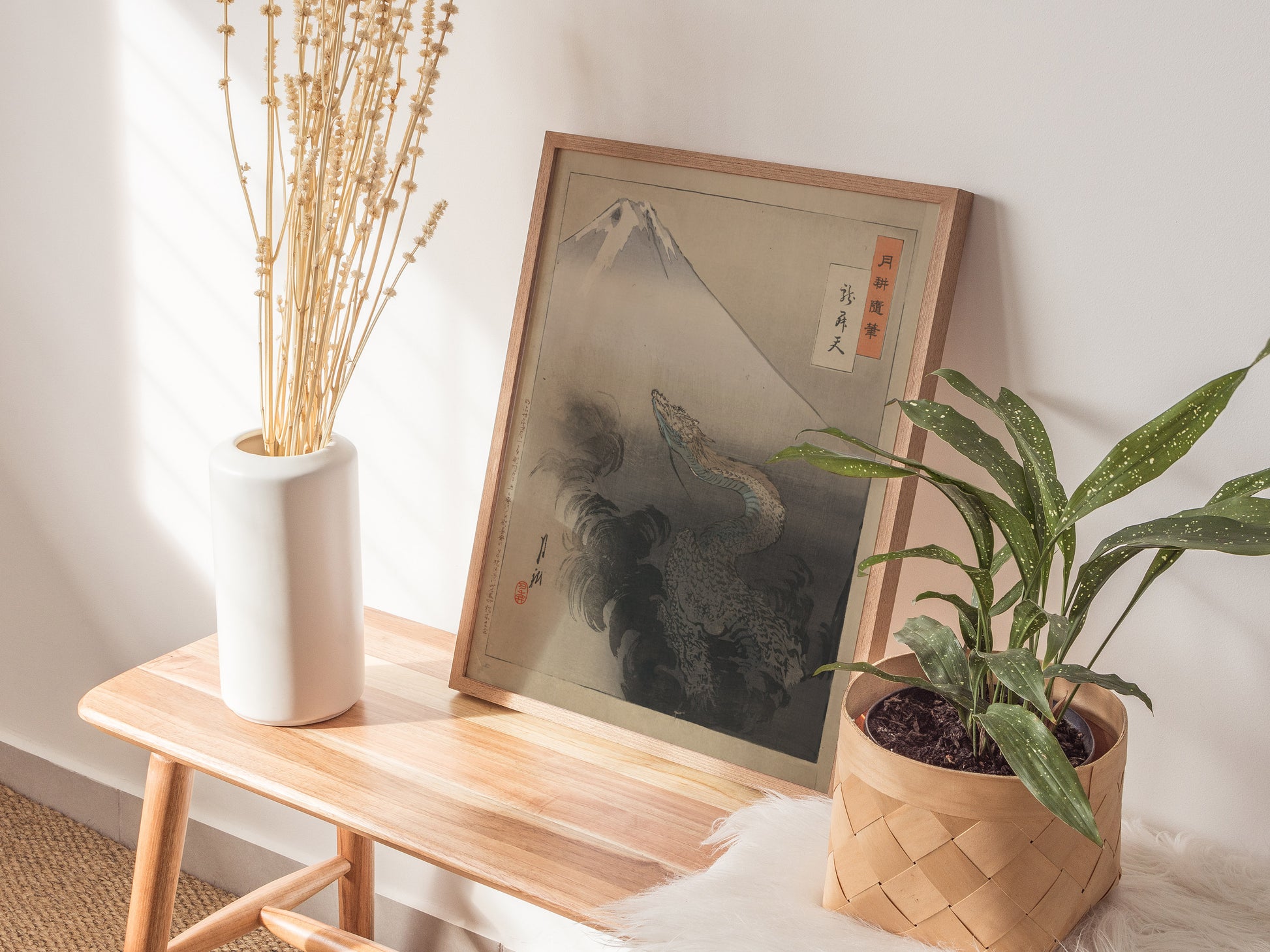 A framed Japanese ukiyo-e print featuring a dragon and Mount Fuji is displayed on a wooden table, flanked by a white vase with dried flowers and a green potted plant.