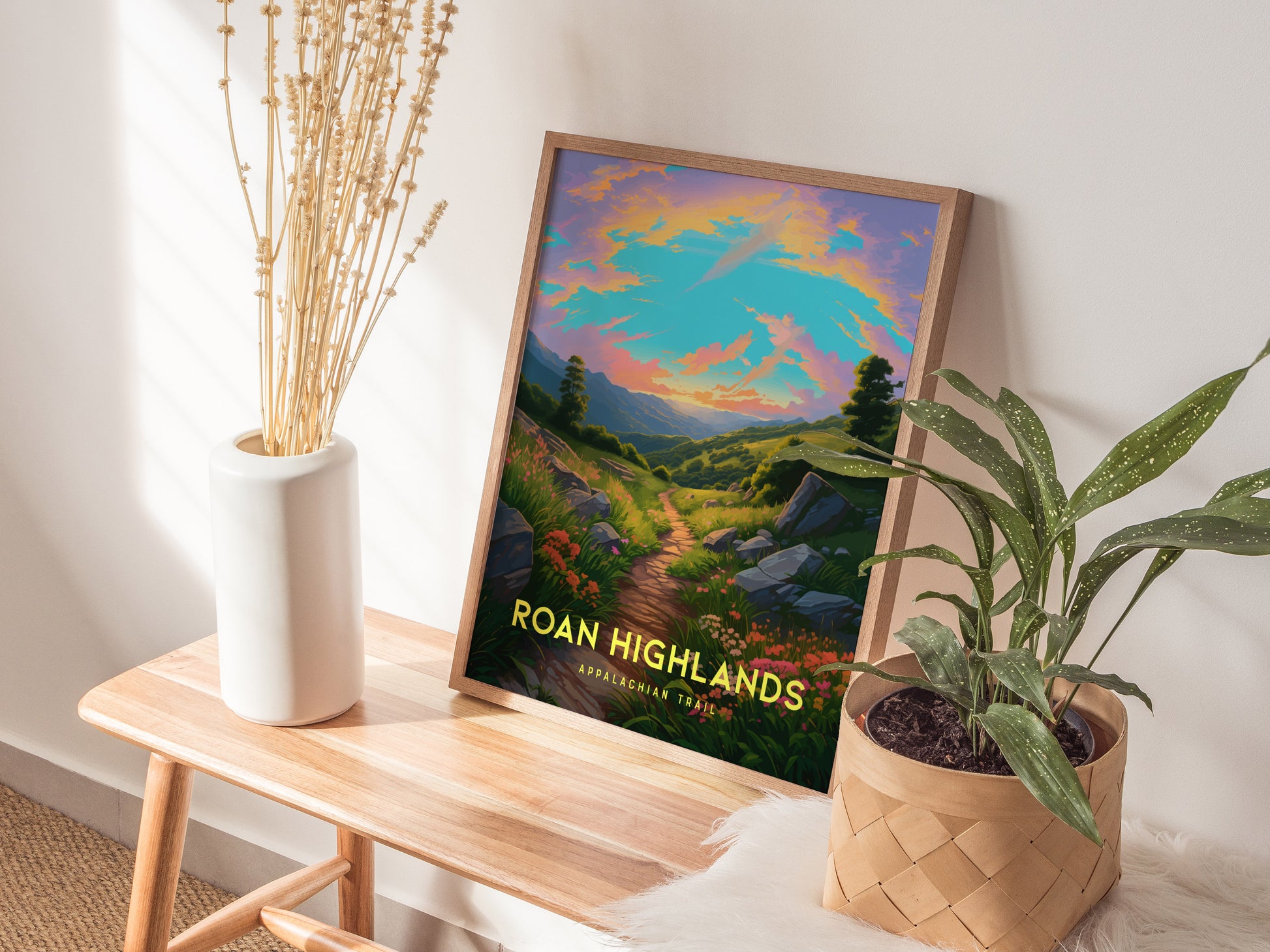 A framed poster of Roan Highlands with a vibrant sunrise over a trail is displayed on a wooden bench, accompanied by a white vase with dried flowers and a potted.