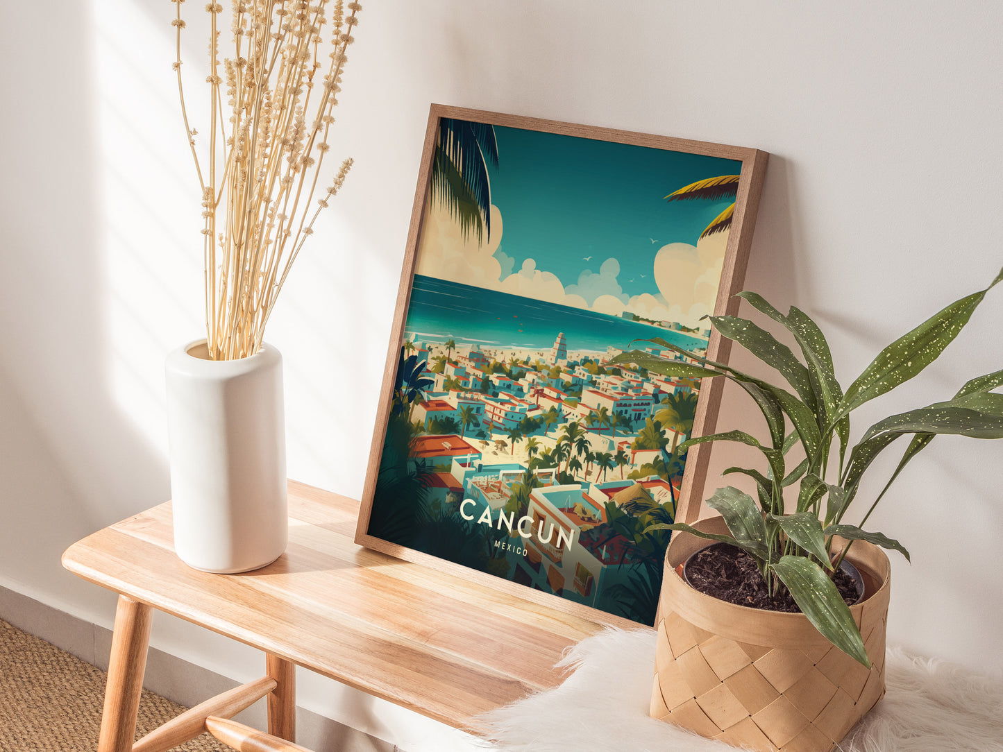 A framed Cancun poster with a vibrant tropical scene rests on a wooden table, accompanied by a white vase with dried flowers and a leafy green plant in a woven.