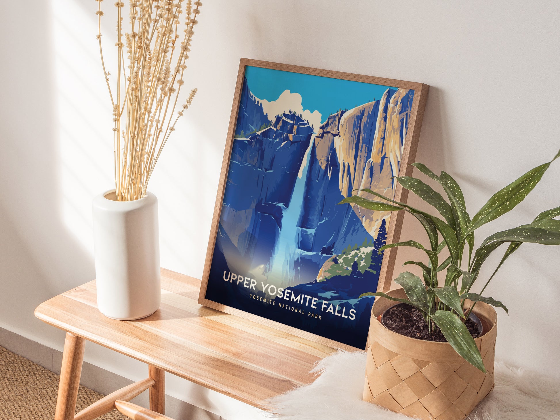 A framed poster of Upper Yosemite Falls is displayed on a wooden bench, flanked by a white vase with dried stems and a potted green plant.