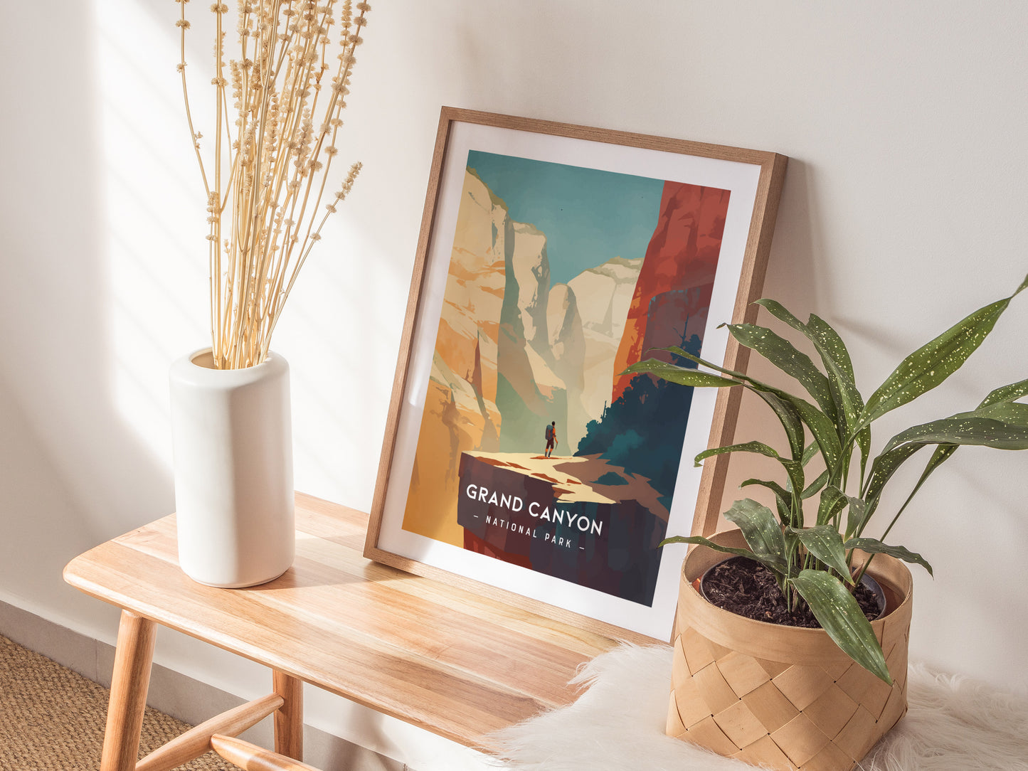 A framed Grand Canyon National Park poster rests on a wooden bench, accompanied by a white vase with dried flowers and a potted green plant.