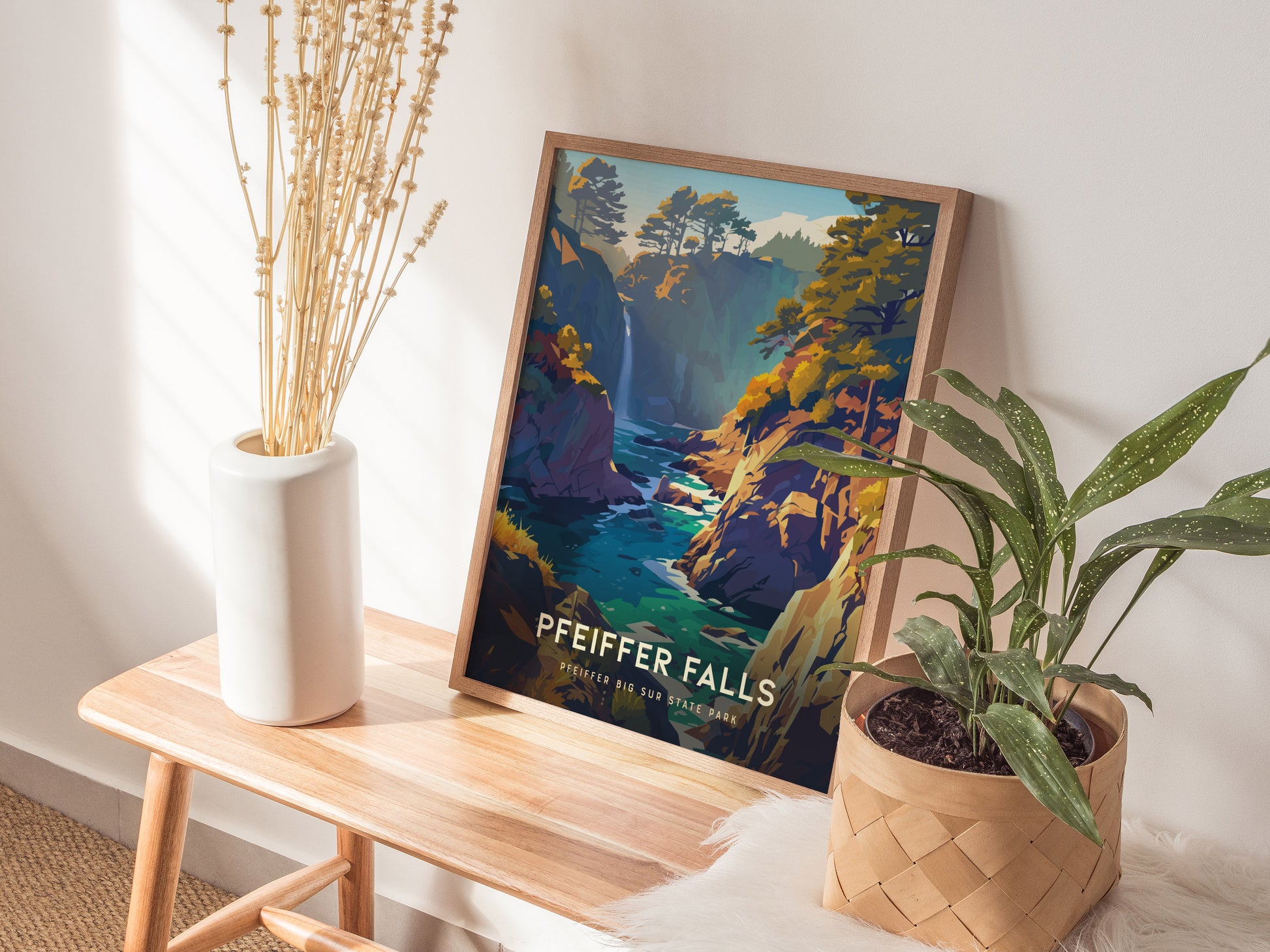 A framed Pfeiffer Falls poster rests on a wooden bench, accompanied by a white vase with dried stems and a potted plant, creating a cozy decor setting.