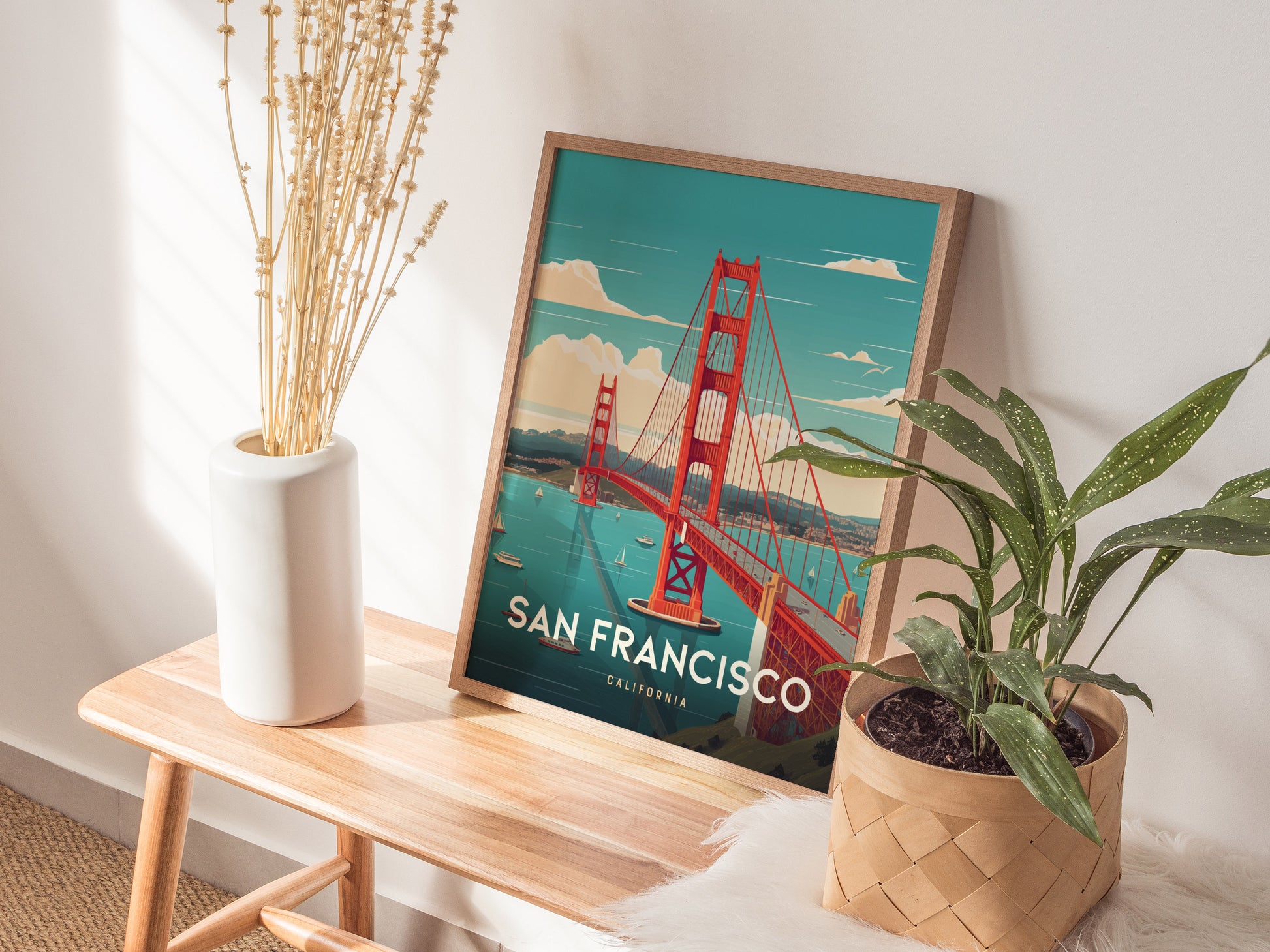A framed San Francisco poster featuring the Golden Gate Bridge rests on a wooden bench, alongside a white vase with dried flowers and a potted green plant.