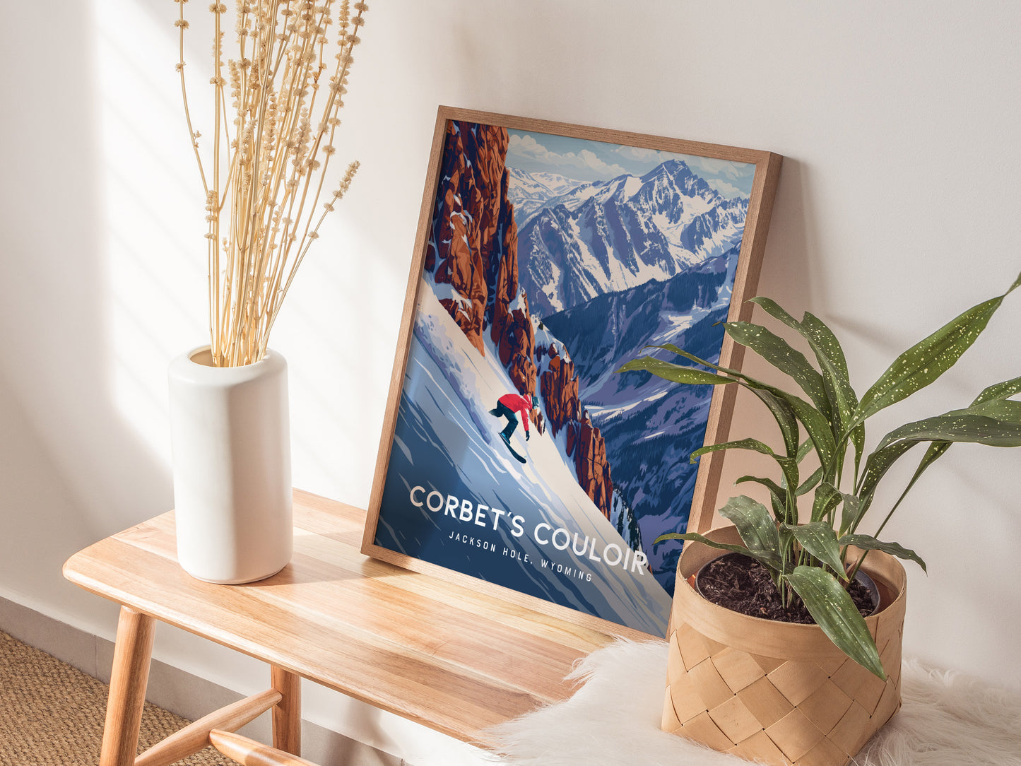 A framed snowboard poster of Corbets Couloir is displayed on a wooden bench, surrounded by a white vase with dried flowers and a green potted plant.