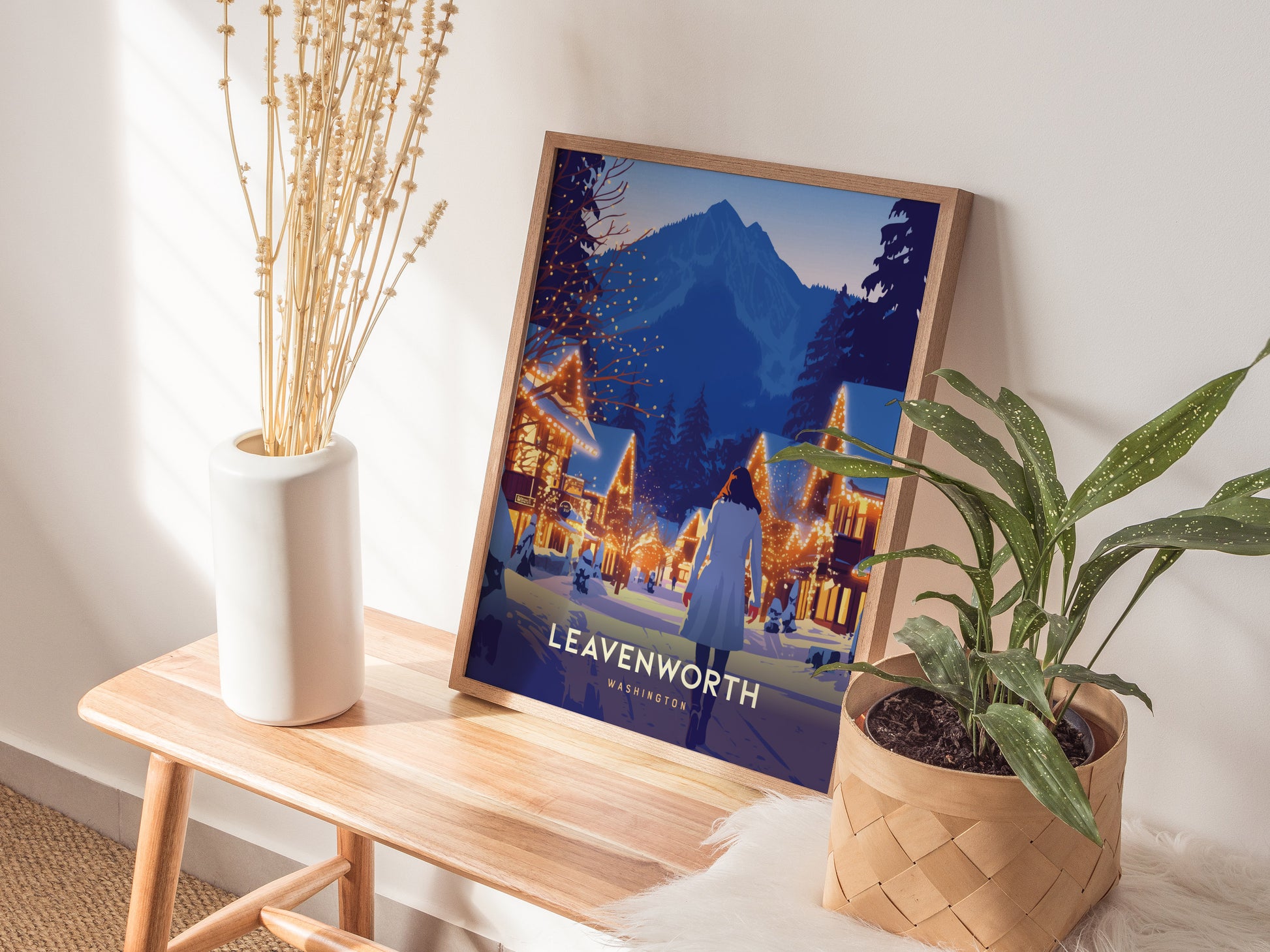A framed Leavenworth, Washington poster rests on a wooden bench, surrounded by a white vase with dried flowers and a potted plant, creating a cozy, natural setting.