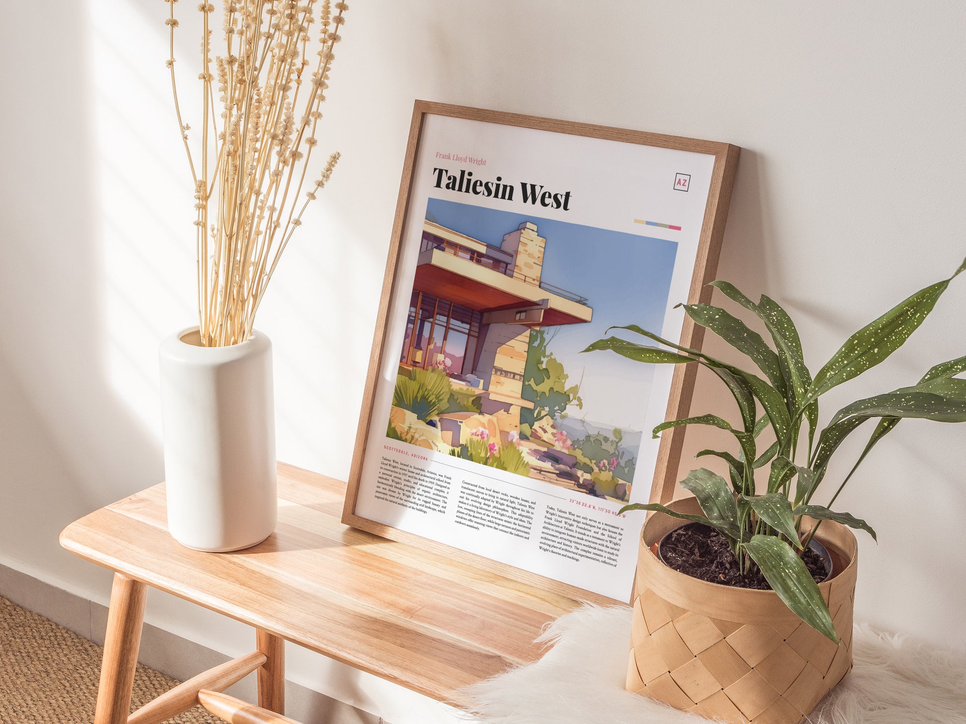 A framed Taliesin West poster rests on a wooden bench beside a white vase with dried flowers and a potted green plant, creating a cozy, sunlit interior scene.
