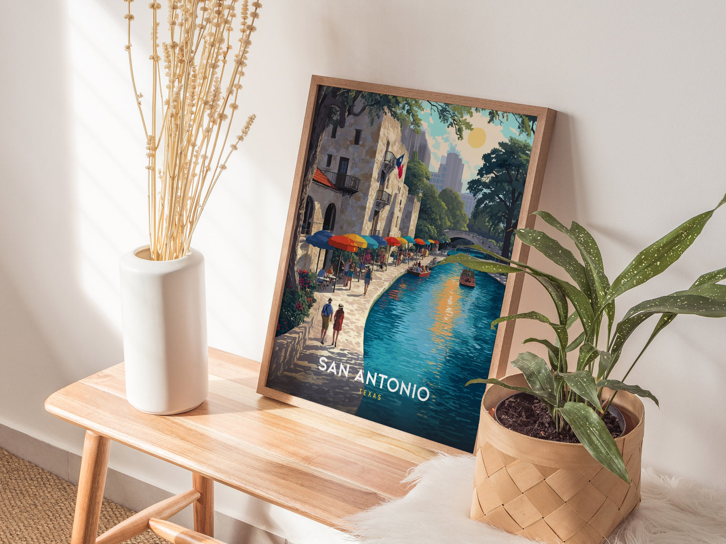 A framed poster of San Antonio, Texas, depicting a colorful riverside scene is placed on a wooden bench alongside a white vase with dried flowers and a potted green plant.
