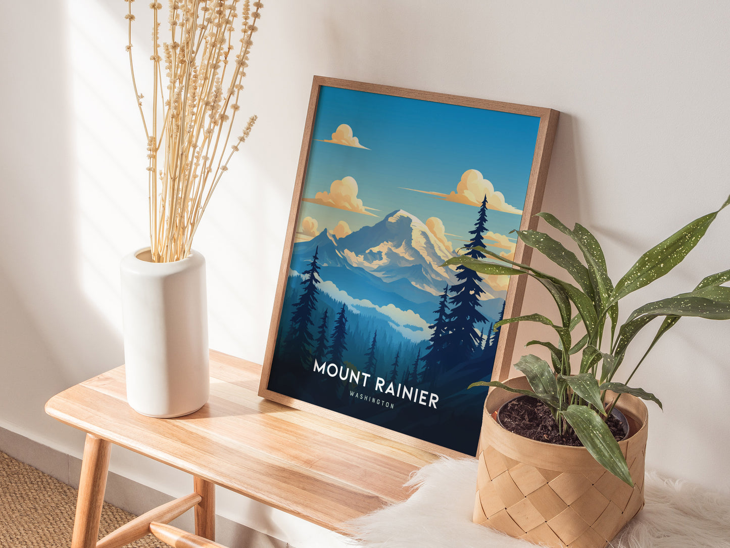A framed Mount Rainier poster leans against a wall on a wooden bench, with a plant in a woven basket and a vase of dried flowers nearby.