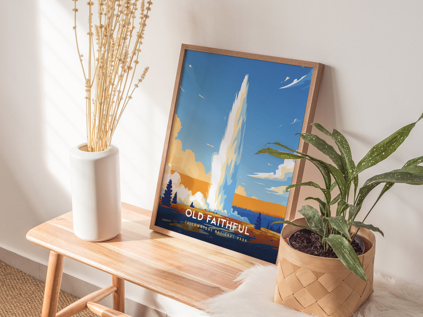 A framed poster of Old Faithful geyser sits on a wooden bench, surrounded by a white vase with dried stems and a potted green plant.