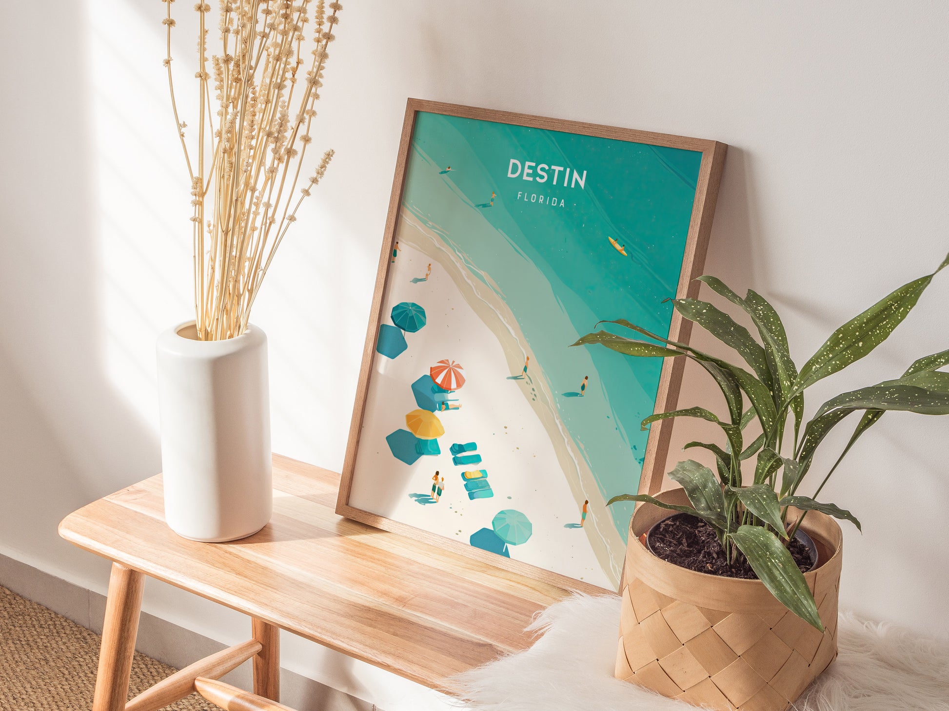 A framed Destin, Florida poster with turquoise waters and beach umbrellas rests on a wooden bench beside a white vase with dried flowers and a potted green plant.