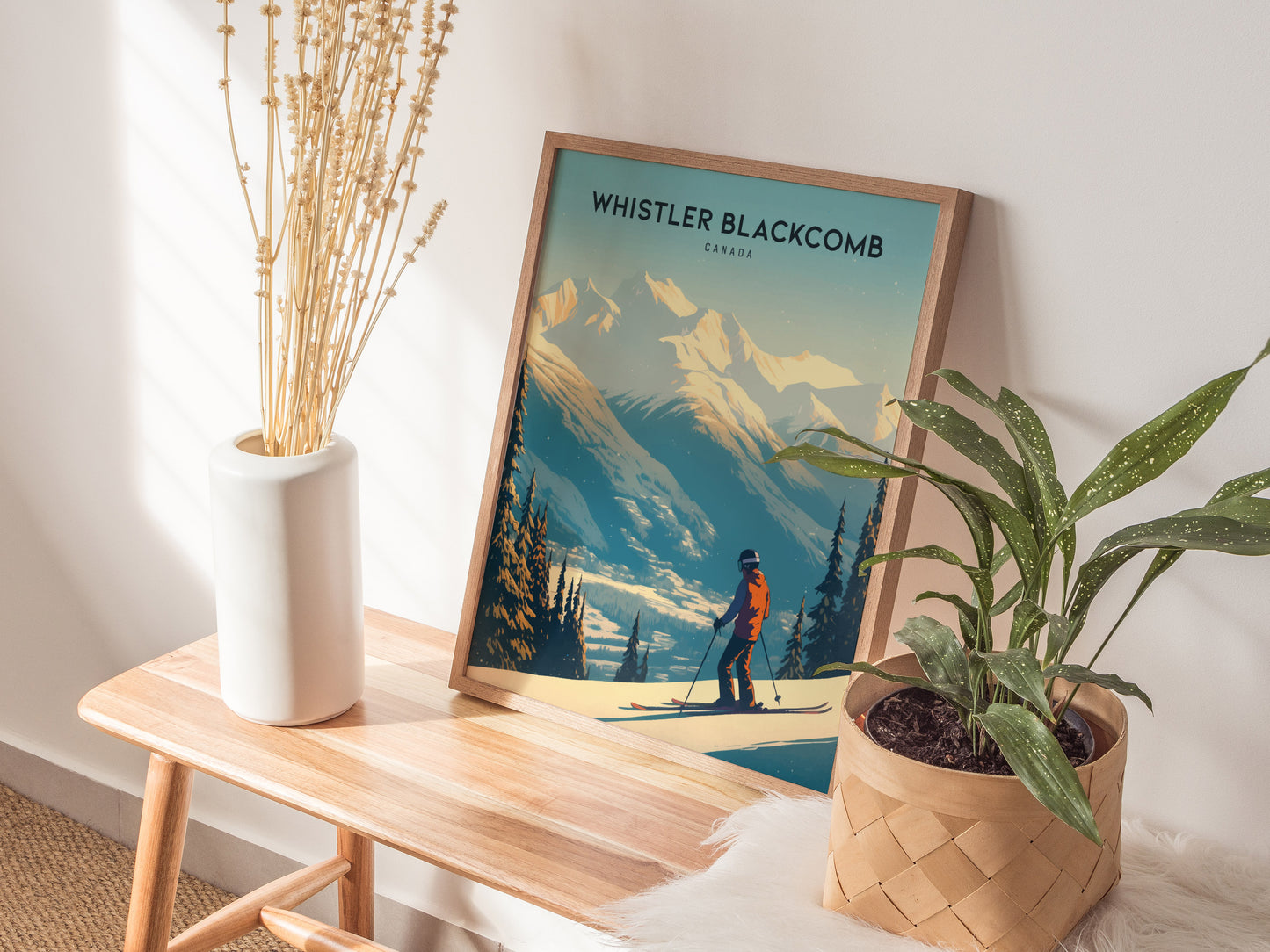 A framed Whistler Blackcomb poster rests on a wooden bench beside a potted plant and a white vase with dried flowers.