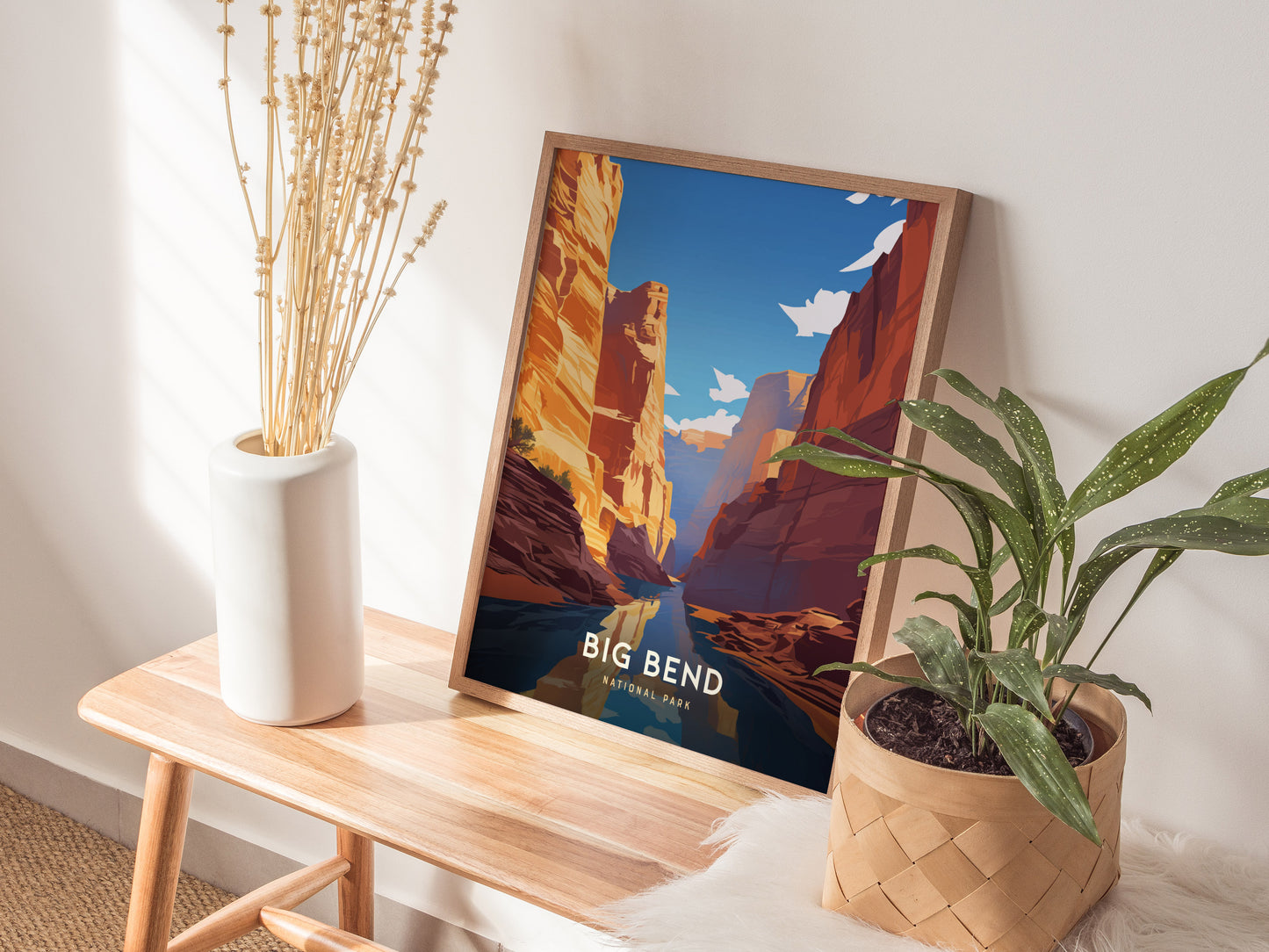 A framed poster of Big Bend National Park leans against a wall, alongside a white vase with dried plants and a leafy green plant in a woven basket.