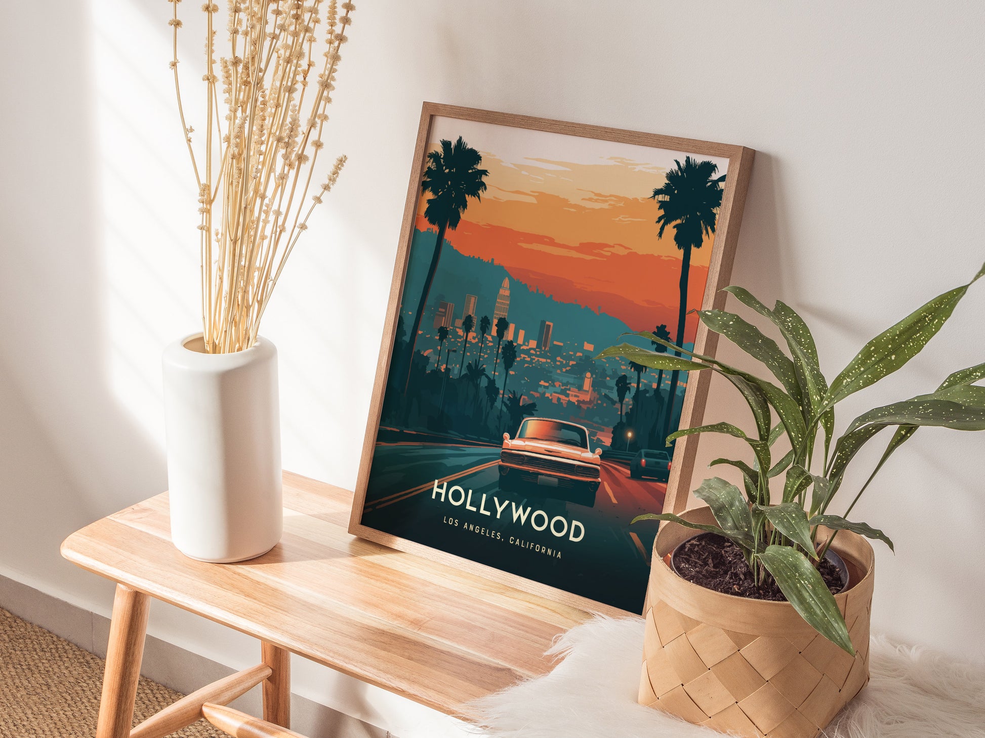 A framed Hollywood poster featuring a sunset view of Los Angeles with palm trees and a classic car is displayed on a wooden bench, accompanied by a vase and potted.