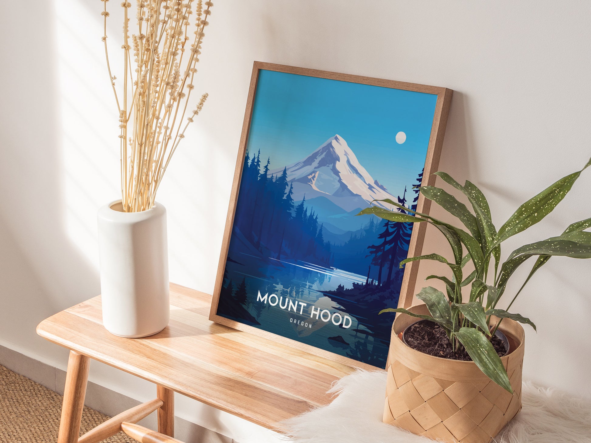 A framed Mount Hood poster sits on a wooden bench, surrounded by a white vase with dried flowers and a potted plant in a woven basket.