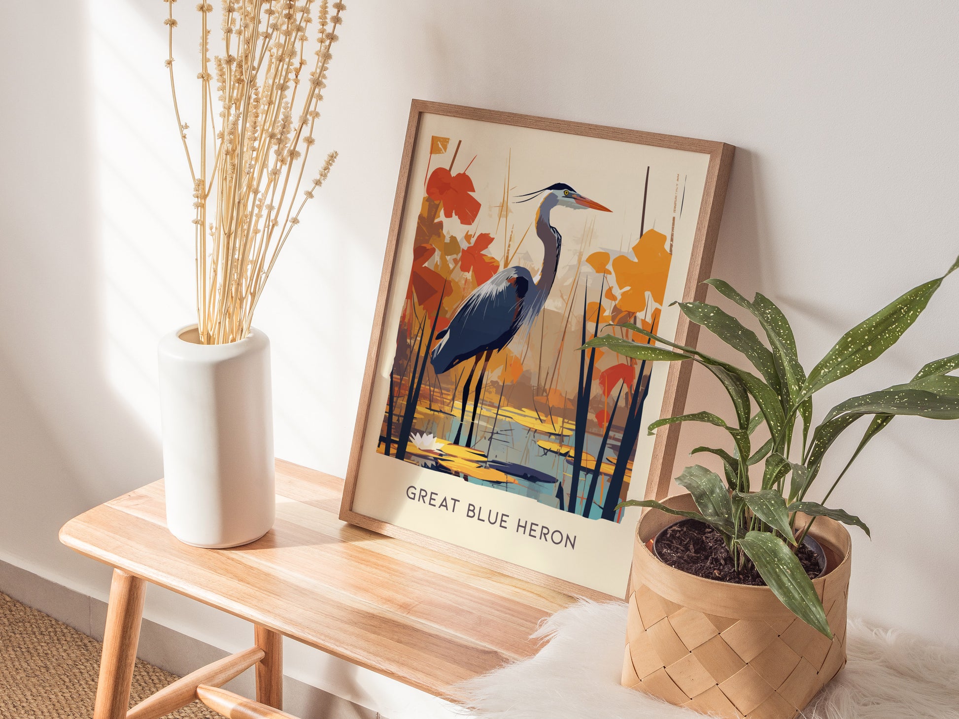 A framed poster of a Great Blue Heron is displayed on a wooden bench, accompanied by a white vase with dried stems and a potted green plant.