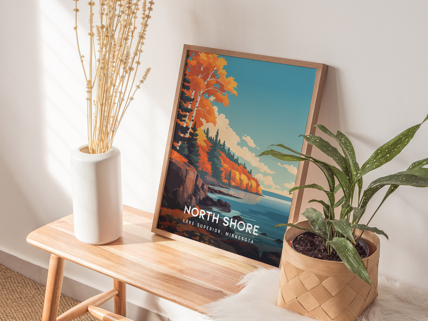 A framed poster titled North Shore, Lake Superior, Minnesota is displayed on a wooden table. The artwork features vibrant autumn foliage along rocky shores, with a tranquil lake and a clear blue sky, complemented by a decorative plant.