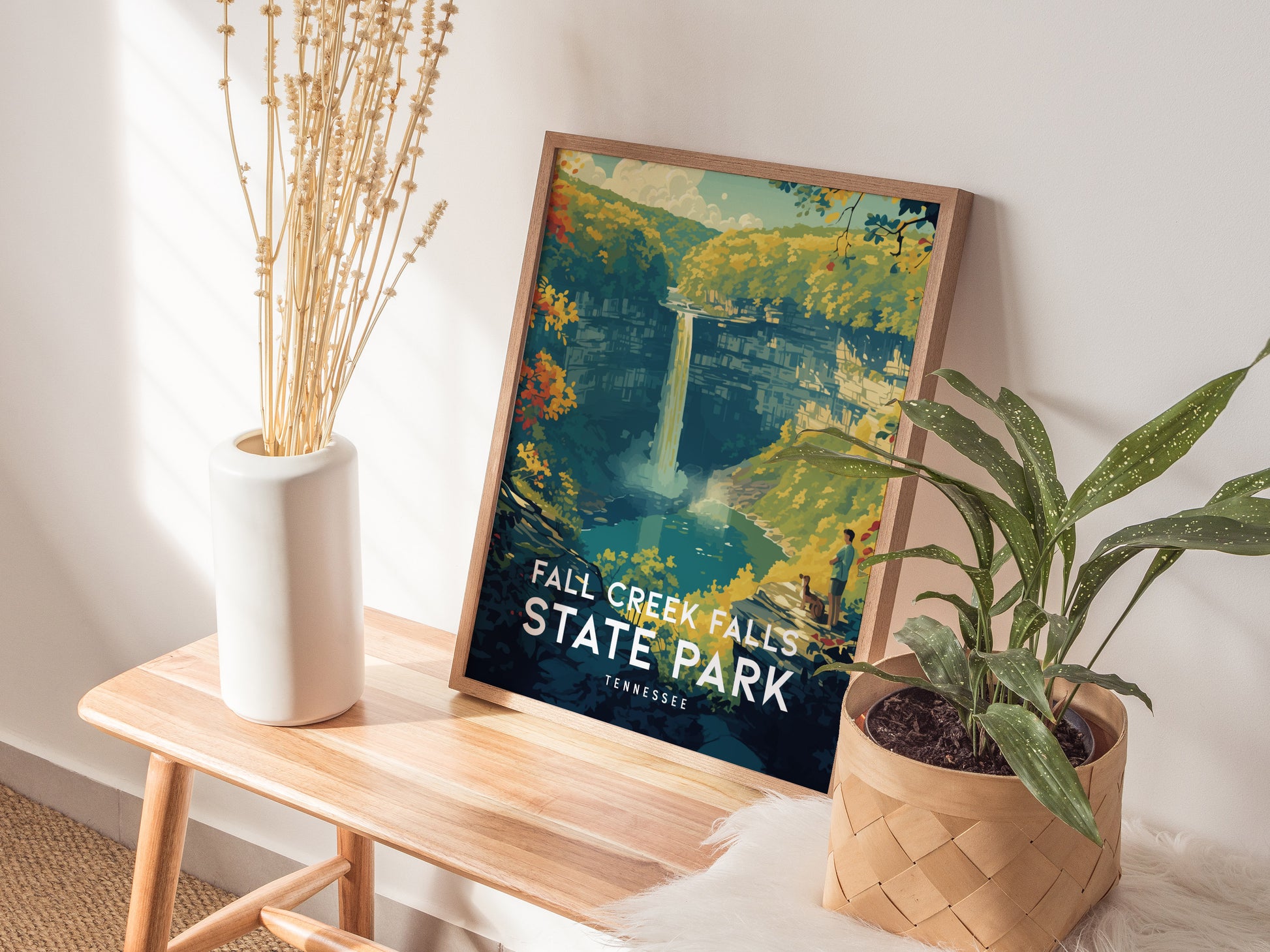 A framed Fall Creek Falls State Park poster rests on a wooden bench beside a white vase with dried plants and a potted green plant, creating a cozy decor setting.